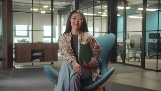 A young asian lady is sitting on the chair in the university, smiling and looking at the camera