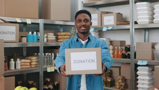 Portrait of positive male volunteer holding cardboard donation box while standing at food bank. African american man in casual clothes smiling and looking at camera.