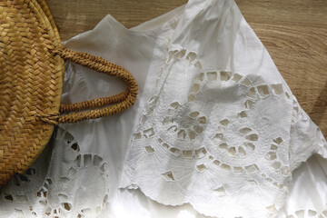 Fototapeta premium White linen lace blouse and round straw bag on wooden background. Flat lay.
