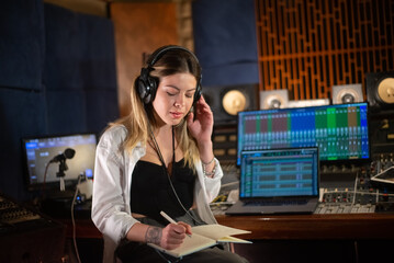 Female music producer listening to music and taking notes in notepad. Young Caucasian woman working in sound recording studio. Creating music concept