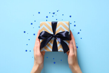 Female hands holding vintage gift box with blue ribbon bow on pastel blue background with confetti....