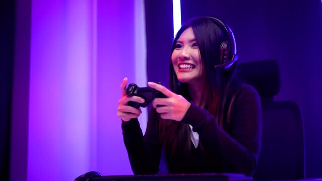Gamer with controller winning victory moment. Young asian pretty woman sitting on chair with computer pc. Female Professional Streamer wearing headphone playing game online in dark room neon light.