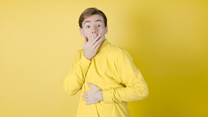 Amazed guy covering his mouth. Looking at camera. Isolated on yellow background. Caucasian guy, 18-20 years old.