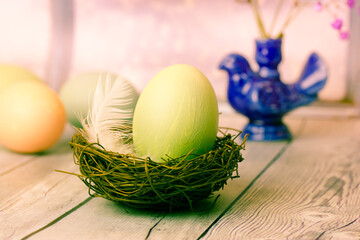 Green painted Easter egg in bird's nest with white feather on wooden table. Rustic Easter background. April holidays postcard. Easter eggs wallpaper. Vase in the form of a blue bird on a background.