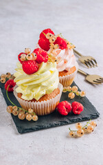 Delicious cupcakes with fresh raspberries and currants on black slate over light beige background.