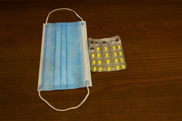 A half-empty blister with yellow tablets and a blue protective mask made of gauze on a wooden table for treatment and recovery