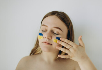 naked portrait of a girl on a white wall. The girl draws the flag of Ukraine on her cheek