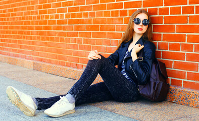 Stylish young woman sitting in the city wearing black rock style over brick wall background
