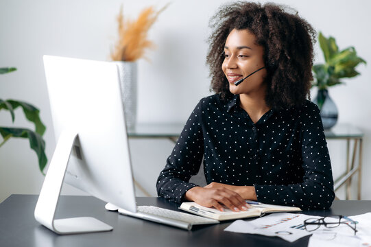 Positive beautiful confident african american woman with headphones, hotline operator, support worker, online consultant sits at computer in modern office, talking to a client on video call, smiling