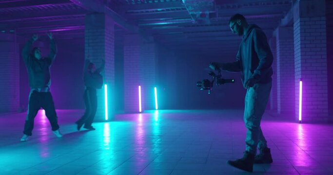 Backstage of video shooting. Cameraman filming hip-hop dancers in neon light. Operator moves with image stabilizer in hands at dark studio. Shooting a clip