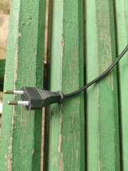 There is a wire on a wooden bench. On a green wooden bench lies a cut piece of black electrical wire with an electrical plug at the end. The wooden sticks of the bench are laid parallel to each other.