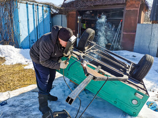 An elderly male welder repairs a car trailer with electric arc welding on a spring sunny day.