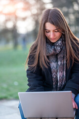 Vertical portrait of young beautiful woman working remotely in a park. She sitting on a bench she is focused as she types on her laptop staring at the keyboard. Remote work outdoors concept.