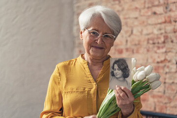 Grandma's Day medium shot of a grandmother wearing sunny yellow shirt holding flowers and black and white photo of her younger version. High quality photo