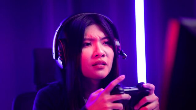 Gamer with controller winning victory moment. Young asian pretty woman sitting on chair with computer pc. Female Professional Streamer wearing headphone playing game online in dark room neon light.