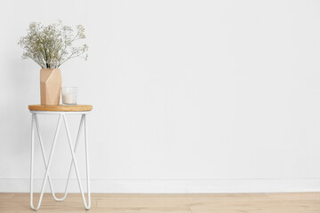Beige vase with gypsophila flowers and candle on table near light wall