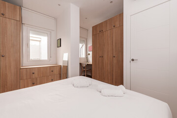 Bedroom with double bed, with white bedding, cabinets with doors in shades of wood, desk with chair and several white aluminum windows