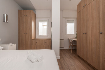 Bedroom with double bed, with white bedding, cabinets with doors in wood tones, desk with chair, chest of drawers, white bedside tables and several white aluminum windows