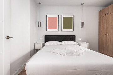 Bedroom with double bed, with white bedding, cabinets with doors in wood tones, black leather upholstered headboard, rolled white towels and white bedside tables