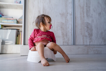 One small caucasian baby girl one year old sitting on the children's potty at home on the floor in day full length with nipple pacifier in mouth front view copy space