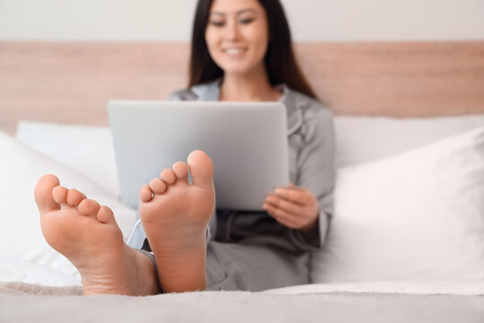 Bare feet of young Asian woman using laptop on bed at home, closeup