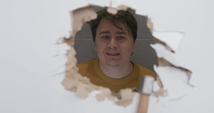 Plump man looks through big hole in white plasterboard wall made with axe. Home owner sits in bathroom against tile smiling behind damage closeup