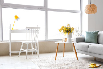 Vase with mimosa flowers on table and sofa in interior of living room