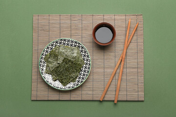 Plate with tasty seaweed sheets, bamboo mat and chopsticks on green background