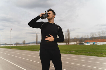A young guy is drinking water and a protein shake after a run on a stadium during the day