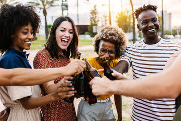 Happy young multiracial friends celebrating and cheering with beer at summer party outdoors - United millennial people laughing having fun together toasting drinks on vacation -