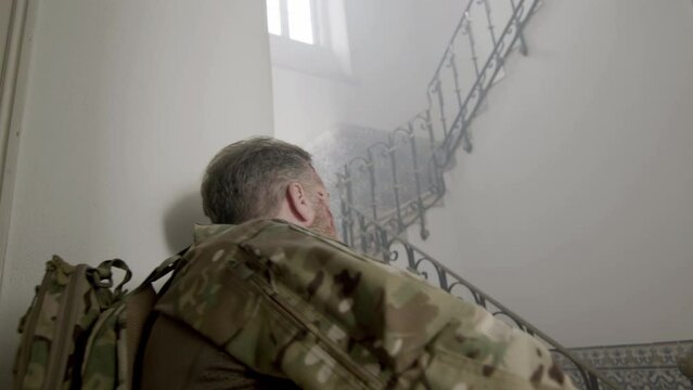 Armed soldier in camouflage uniform with rifles climbing ladder. Medium shot of focused msn with scratches on face looking up, storming hostile building. Assault, battle, attack concept