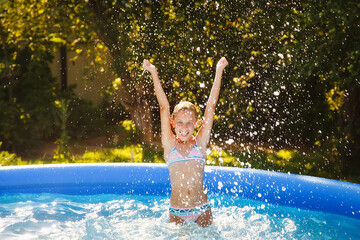 The child jumps out of the water. A girl splashes in an inflatable pool in the garden on a sunny summer day.