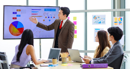 Asian professional successful businessman manager standing showing company growth profit target graph chart presentation on computer monitor to young male and female colleagues in office meeting room