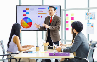 Asian professional successful businessman manager standing showing company growth profit target graph chart presentation on computer monitor to young male and female colleagues in office meeting room