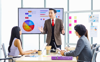 Asian professional successful businessman manager standing showing company growth profit target graph chart presentation on computer monitor to young male and female colleagues in office meeting room