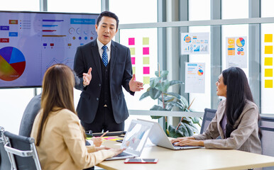 Asian professional successful businessman manager standing showing company growth profit target graph chart presentation on computer monitor to young male and female colleagues in office meeting room