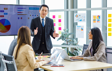 Asian professional successful businessman manager standing showing company growth profit target graph chart presentation on computer monitor to young male and female colleagues in office meeting room