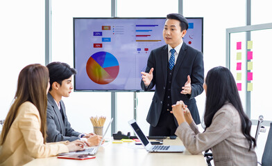 Asian professional successful businessman manager standing showing company growth profit target graph chart presentation on computer monitor to young male and female colleagues in office meeting room