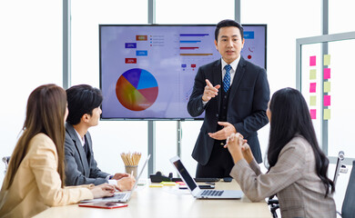 Asian professional successful businessman manager standing showing company growth profit target graph chart presentation on computer monitor to young male and female colleagues in office meeting room