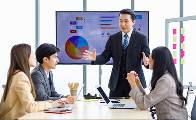 Asian professional successful businessman manager standing showing company growth profit target graph chart presentation on computer monitor to young male and female colleagues in office meeting room