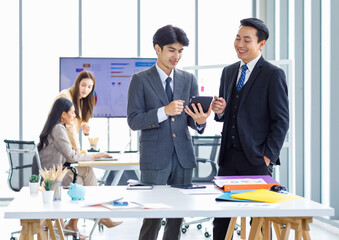Asian male professional businessmen colleague in formal business suit take coffee break standing holding disposable paper cup and tablet computer talking have conversation together in front monitor