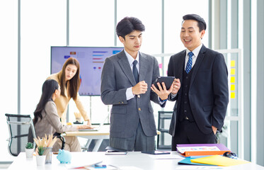 Asian male professional businessmen colleague in formal business suit take coffee break standing holding disposable paper cup and tablet computer talking have conversation together in front monitor