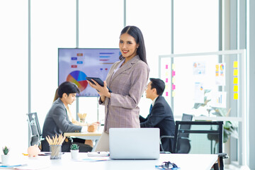 Millennial Asian young happy beautiful female businesswoman employee staff in formal business suit standing smiling holding touchscreen tablet computer in hands posing in company office meeting room