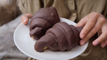 Two croissants in chocolate glaze on a white plate are held by a female waitress, taking one croissant to serve on the table for breakfast. Chocolate croissants with a high content of sugar and cocoa.