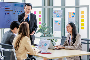 Asian professional successful businessman manager standing showing company growth profit target graph chart presentation on computer monitor to young male and female colleagues in office meeting room