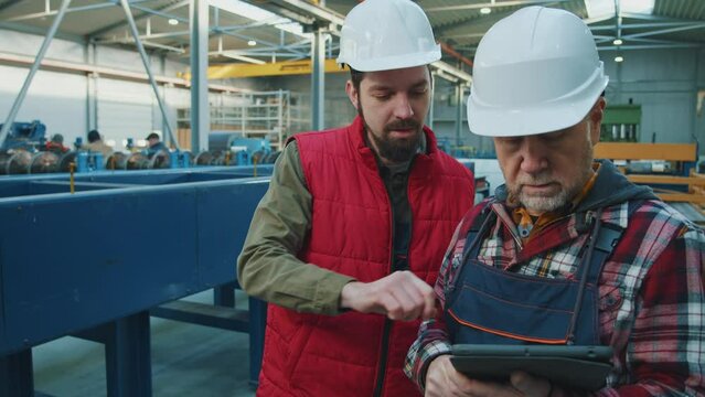 Young and elder engineers men heavy industrial workers with helmet using a digital tablet in factory. Checks production processes. Inspector in warehouse.