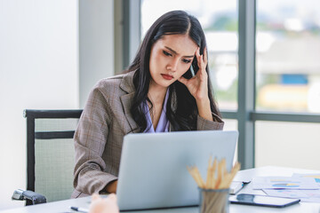 Asian young unhappy upset stressed depressed frowning face female businesswoman employee staff in formal business suit sitting at working desk holding hands on head having problem trouble headache
