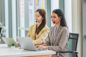 Two millennial Asian young beautiful female customer service call center in formal business suit wearing microphone headset sitting working talking with customers at working desk in company office