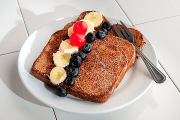 breakfast with french toast with sugar and cinnamon and slices of banana, blackberries and cherries on top, on a white plate and a cup of hot creamy coffee on the side