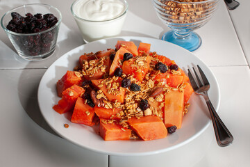 A breakfast with papaya granola and blackberries on a white plate and on a white table with a small cup of yogurt, granola and blackberries on the side, side view shot.